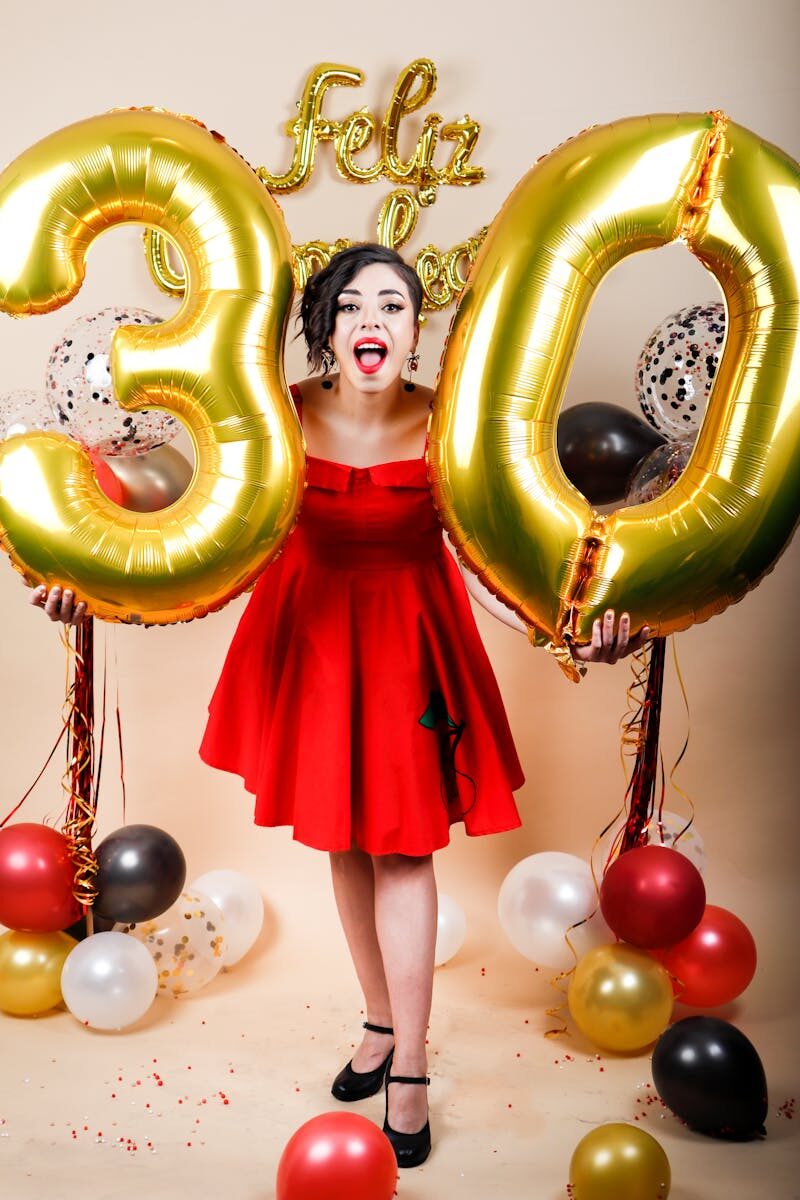 Woman in red dress celebrates 30th birthday with gold balloons and festive decor indoors.