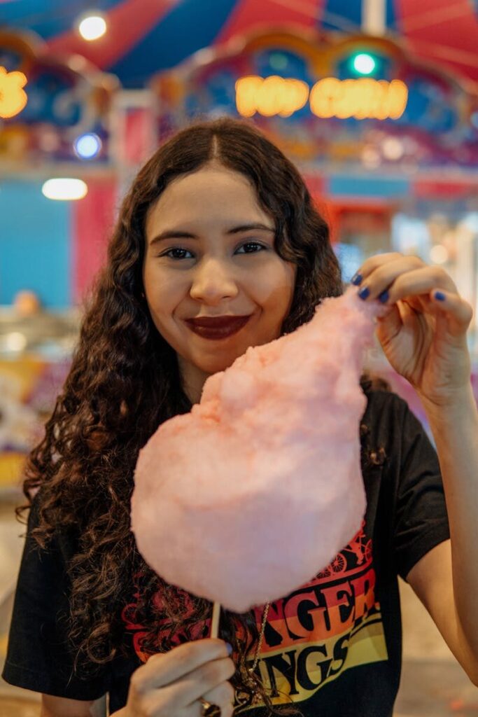 Smiling woman holding pink cotton candy at a vibrant fair setting.